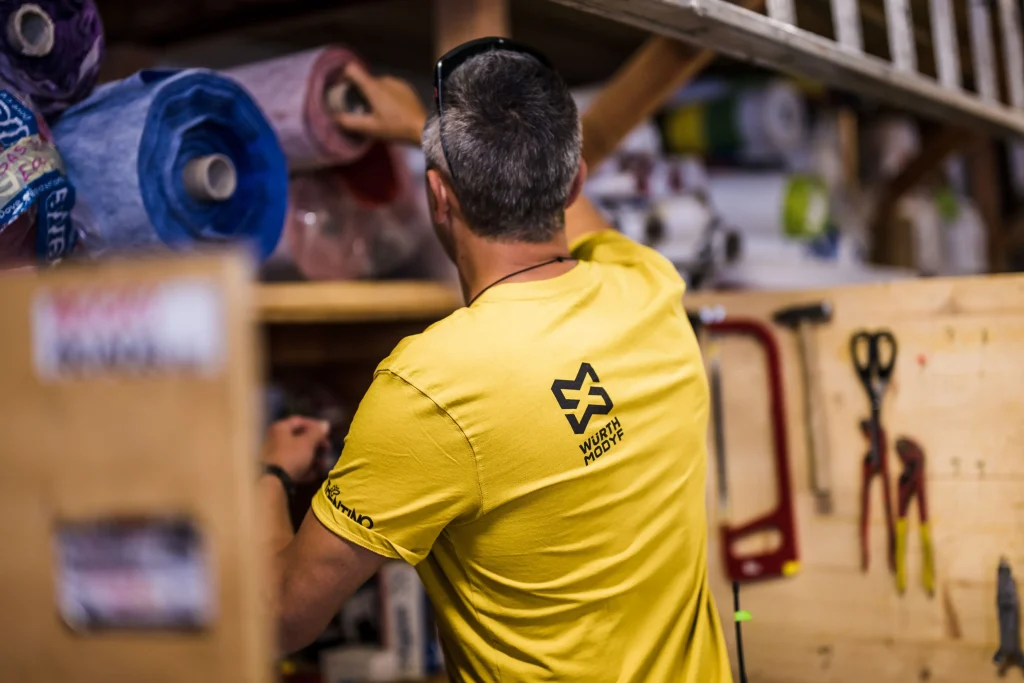 Un homme en t-shirt de travail jaune est de dos, fouille dans une étagère