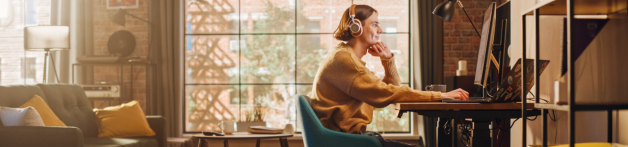 Une jeune femme est chez elle, devant son écran d'odinateur avec un casque sans fil sur la tête et navigue sur internet