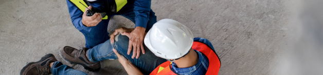 Un ouvrier portant un casque blanc et un gilet orange vient soigner un autre ouvrier qui s'est fait mal au genou en tombant
