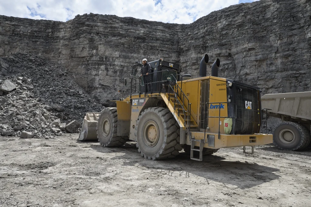 Un homme en tenue de travail se trouve sur un chantier, il se tient sur une grande pelleteuse jaune