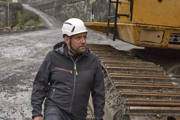 Un homme en tenue de travail se trouve sur un chantier boueux, il se tient à coté d'une machine de chantier jaune