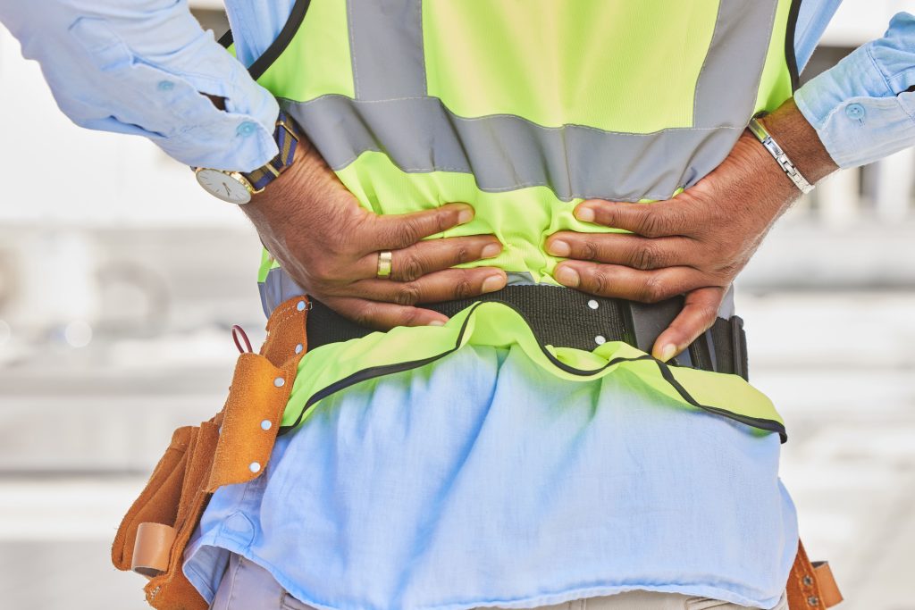 Un ouvrier se tient le dos de douleur. Il porte une chemise bleue ciel, avec par dessus un gilet de signalisation haute visibilité jaune. Il porte une ceinture d'outils.