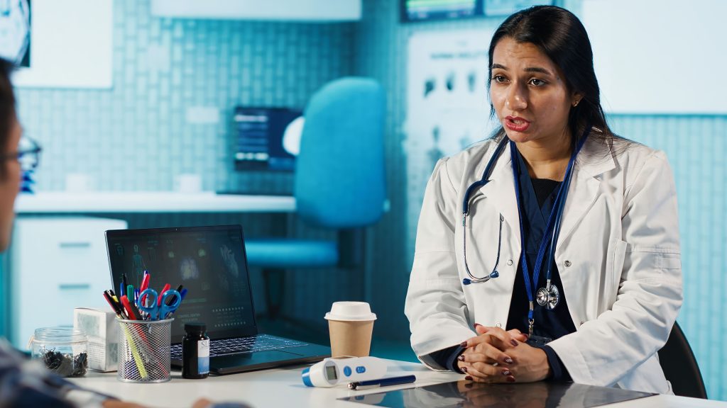 Une femme médecin est en pleine consultation avec une patiente, à son bureau. Elle porte un stéthoscope ainsi qu'une blouse blanche. Elle se trouve dans son cabinet médical.