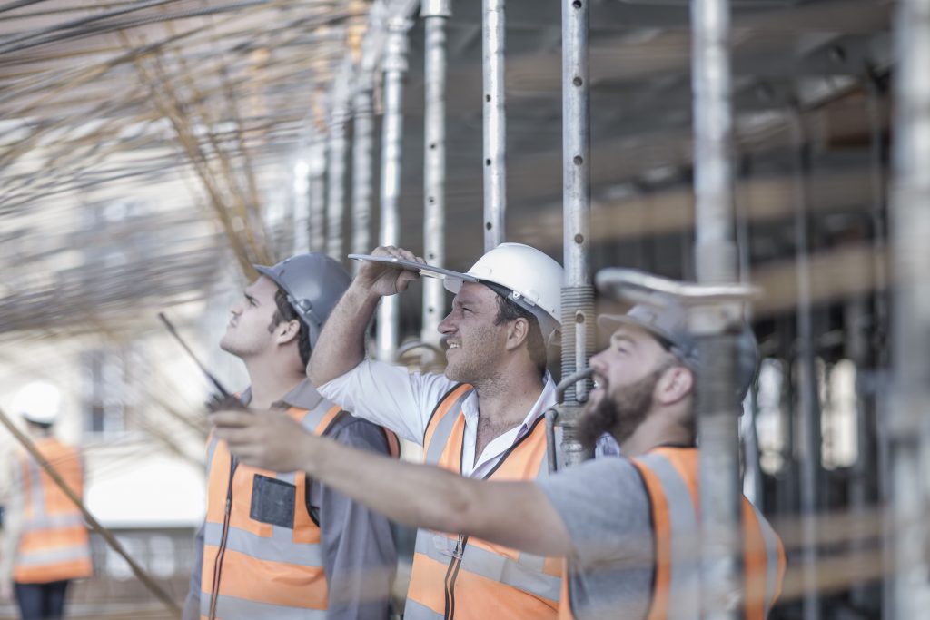 Trois ouvriers avec des casques blancs et gris et des gilets haute visibilité oranges regarde et pointe du doigt quelque chose au dessus d'eux