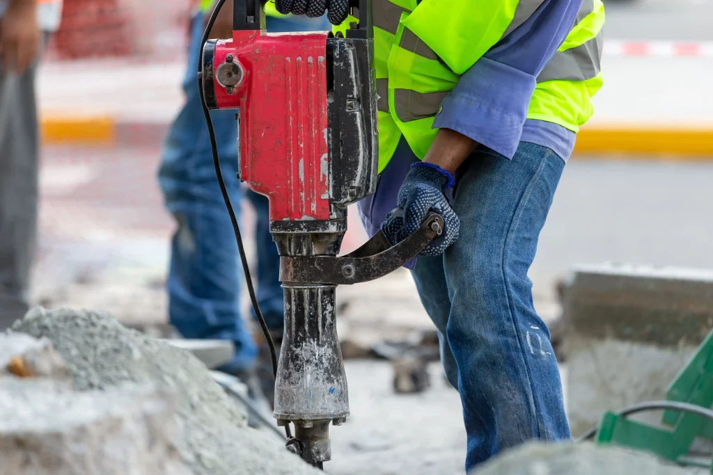 Un homme en tenue de chantier haute visibilité utilise un marteau piqueur pour casser un sol.